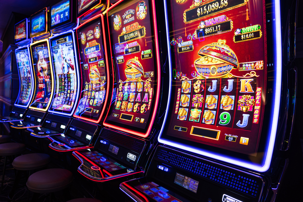 A row of brightly lit, modern slot machines in a casino, with glowing screens displaying jackpots and symbols